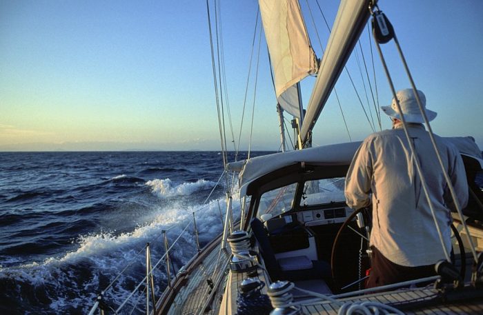 A landscape (layout) photo taken from the cockpit of a sailboat under sail in the Gulf of Chiriqui, on Panama’s Pacific coast. The horizon bisects the image horizontally, balanced with blue sky above and blue ocean below. Golden clouds can be seen on the left edge of the photo from an off-camera sunset. The forward half of a sailboat’s cockpit, mast, and bow is visible on the right side of the image. Its boom stretches from overhead to the mast in the center of the image, where it intersects with the horizon. A man steers at the helm as the boat plunges into a white foam wave off the forward port quarter, which points the bow towards the horizon line.