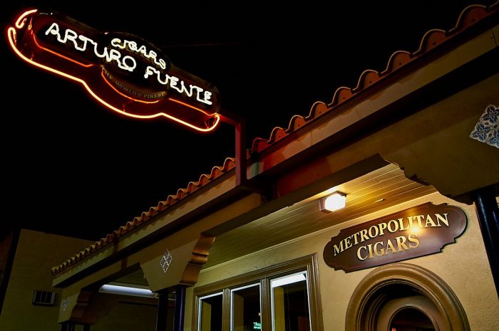 Landscape photo of the Arturo Fuente cigar store in Ybor City, at night. The store front’s Mediterranean tiled roofline angles from the upper right corner down to the lower left corner of the image. In the upper left corner of the image, an oval-shaped neon sign angles 45-degrees out from the tiled roof. The roofline and sign form an X, framed by a dark black sky. The sign proclaims: “Cigars, Arturo Fuente” in yellow neon letters, surrounded by a red neon border. In the lower right third of the image an oval varnished wooden sign states “Metropolitan Cigars” in yellow letters over the top of a rounded, arched wooden door frame.