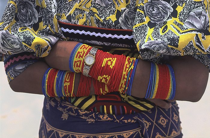 A close-up landscape photo of a Kuna woman in the Guna Yala region of Panama. The top of the frame starts just below her shoulders and continues several inches below her waist. Arms folded at her waist, each arm is wrapped with a single strand of colorful beads, with clusters of blue, red, and yellow sections. A watch is strapped over the wrist of her topmost arm. Her yellow print blouse displays large gray flowers outlined in black, with black leaves surrounding the buds. Her dark blue skirt features large beige-colored abstract patterns. She is a showcase of multiple colors.