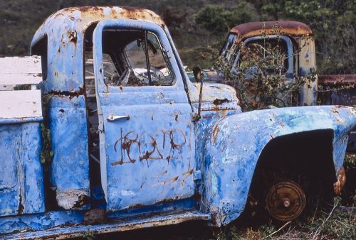 Horizontal photo of a 1940s vintage pickup truck that has been abandoned on the island of Maui, Hawaii. The photo was taken from the right side of the truck, with the front of the truck pointing to the image’s right. The truck’s bulbous rounded cab appears just large enough for two people. The right front tire has been removed underneath the fender. A rusted wheel sits directly on the ground. The truck’s patchy blue paint—light sky blue to a deep azure—makes the truck pop against the dark brown and green hillside in the background. Dark brown rust spots punctuate and accent the blue exterior. In the top right corner of the photo the cab of another abandoned truck, visible over the blue truck’s fender, faces the opposite direction—as if two lonely friends sit silently together.