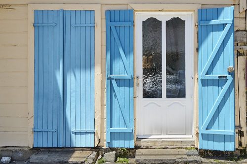 Horizontal photo of a white door with full-length blue shutters on either side in coastal town of Grand-Bourg on Marie-Galante Island in the Caribbean. Two full length panes of rippled glass divide the white door, which appears in the right side of the image. The long shutters on either side of the door are built with narrow planks of wood, framed with two Z-shaped supports that zig zag across the inside of the shutters. The Z-shaped pieces of wood cast deep shadows in the overhead sun. To the left of the door and its open shutters we see another set of blue shutters which are closed. The exterior wall has been painted a light beige.