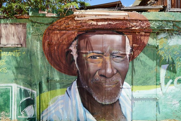 Horizontal photo of an artist’s mural painted on the side of a building in the town of Grand-Bourg on the island of Marie-Galante. The mural is a portrait of a black man who appears to look directly at the camera’s lens. He squints into the direct sun, lips drawn tightly, giving the appearance of a mild grimace. White hair, white eyebrows, and white beard stubble frame his long face underneath a reddish-brown straw hat which is folded up above his forehead. His leading right shoulder dips forward slightly. An open shirt, with large light blue and white vertical stripes, opens to his black-skinned chest. Multiple colors of green: pastel teal, lime, mint, olive, and pine, surround his head and shoulders. A metal door comprises most of the building wall. Hinges, strips of metal brackets, and rivets define the mural’s background texture. At the top of the photo weathered 2x4s and a rusted corrugated roof protrude above the top of the building.