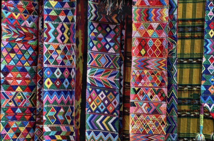 A close-up photo (landscape layout) of colorful cloth belts hanging vertically at the Chichicastenango market, Guatemala. The embroidery on each belt is extremely finely stitched, showing off significant detail in each piece. Patterns include diamonds, zigzags, Xs, chevrons, stripes, and more. Most of the belts are multicolored—with every color imaginable in the color wheel.