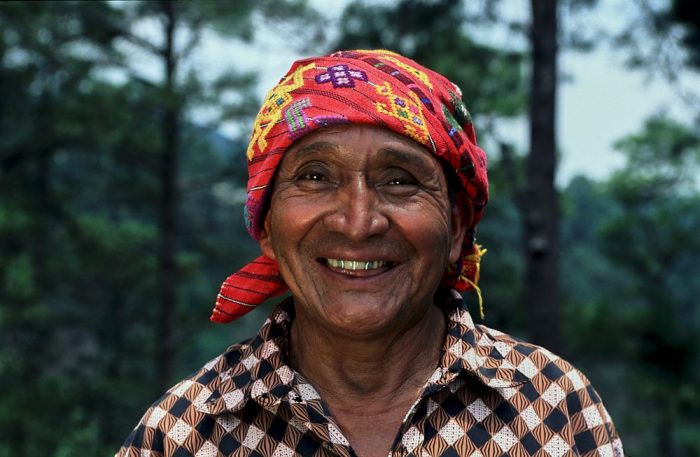 Close-up landscape photo head and shoulder portrait of a Guatemalan shaman on a mountainside in Chichicastenango. He is centered in the shot, with blurred dark green trees in the background on the left and right third of the image. The light is soft and diffuse. His shirt prominently displays a 1-inch diagonal checkered print with an unusual combination of colors: black, white, and light coral. He wears an embroidered red headscarf, with accents of yellow and purple, tied at the back of his neck. He smiles broadly at the camera, showing off two silver capped upper teeth.