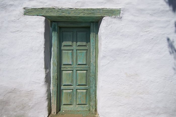 A horizontal landscape photo of a white stucco wall in Old Town, San Diego. The wall is brightly illuminated by the full-on overhead sun. A vertical door, just left of center, is framed with weathered wood painted shades of olive and teal green. Above the door a six-foot long lintel is embedded into the stucco wall. Eight rectangular panels, carved into the door, cast shadows below each panel. The olive-teal colored lintel and door contrast dramatically against the white stucco wall. At the right side of the image, shadows from a nearby tree frame the edge of the photo in a narrow strip along the vertical length of the white wall.