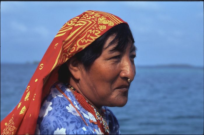 Landscape portrait of a Kuna woman in the Guna Yala region of Panama. We see the right side of her face, which is exposed to the sun, as she gazes off-camera to the right. She smiles, but with her mouth closed. A gold nugget, which glints in the sunlight, pierces the lower part of her nose. On her head she wears a long red cotton scarf, with yellow accents, which trails off her shoulders and below the image frame on the lower left. Bangs hang down from underneath the scarf and sweep across her forehead. She also wears a light blue cotton blouse with white and darker blue floral prints—and a necklace of tiny red beads looped into multiple stranded layers. Behind her a blurred ocean and sky forms a solid blue in the distance.