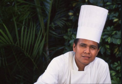 This landscape portrait of a chef in San Salvador, El Salvador, shows him in a double-breasted, white chef’s uniform. He is a young, handsome El Salvadoran man in his late twenties. He faces the camera directly. In the bottom of the frame, we see his torso just above his elbows. His right shoulder is positioned just below center in the image. He leans to his left, such that his face is in the right 1/3 of the frame. He wears a 2-foot-high white chef’s hat that leans upward and to the right at a 25-degree angle. Dark green tropical foliage blurs in the background on the left side and right edge of the photo, making his white uniform and conspicuously tall hat pop in the foreground.