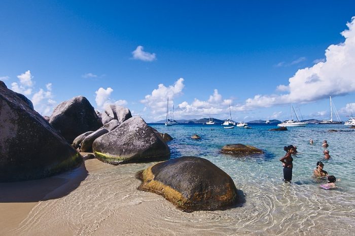 Deep blue sky frames the upper half of this horizontal landscape photo of The Baths in the British Virgin Islands. White cumulus clouds arc from the upper left, down across the horizon, and up again to the right. Beige sand fills the lower third of the image, while large boulders cascade from the left into the center. Crystal clear waters lap against the boulders and ripple out into the turquoise waters of Devil’s Bay. Families swim in the shallow waters on the right side of the photo. Ten or more sailboats drift at anchor on the horizon, several hundred feet in the distance.