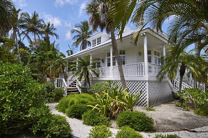 Landscape photo of a home on Useppa Island, Florida. The image was taken from the right and front of the house, at about a 30-degree angle and 40-feet distant. The white house—in the Key West architectural style—has an elevated porch which wraps around on all four sides. A small, windowed dormer can be seen in the center of a silver metal roof. The front stairs descend from the porch to a manicured pea-gravel walkway that extends toward the bottom of the image. Lush tropical palms and foliage in multiple shades of green frame all sides of the house, with blue sky and small wisps of white clouds overhead.