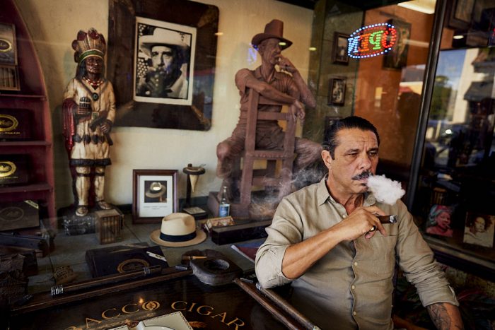 A horizontal photo of Pedro Berro, owner of Cuban Tobacco Cigar Company, who sits in front of his store in Little Havana, Miami. As he smokes one of his cigars, his right elbow rests on a square table with the name of the store visible on the surface. His right hand holds a dark, fat, ash-laden, lit cigar inches below his chin. He stares at a 45-degree angle toward the right side of the image as he exhales a small cloud of dense white smoke. Behind him, through the glass window of the storefront, visible above Pedro and to the left, a full-size wood carving of a man sits facing backwards on a simple chair. The man rests his right arm on the top of the chair as his left arm, bent at his left elbow, holds a wooden cigar to his mouth. The statue’s pose, actions, and direction of his gaze almost perfectly mirror Pedro. We see additional cigar-related paraphernalia in the window adjacent to the statue: a B&W photograph of another man smoking, what appears to be the straw hat of the man in the photo on the surface below, and another, smaller, wooden statue of an Indian chief with full headdress holding a cylindrical collection of cigars in his right hand. The Indian stares off in the same direction as Pedro and the other statue.