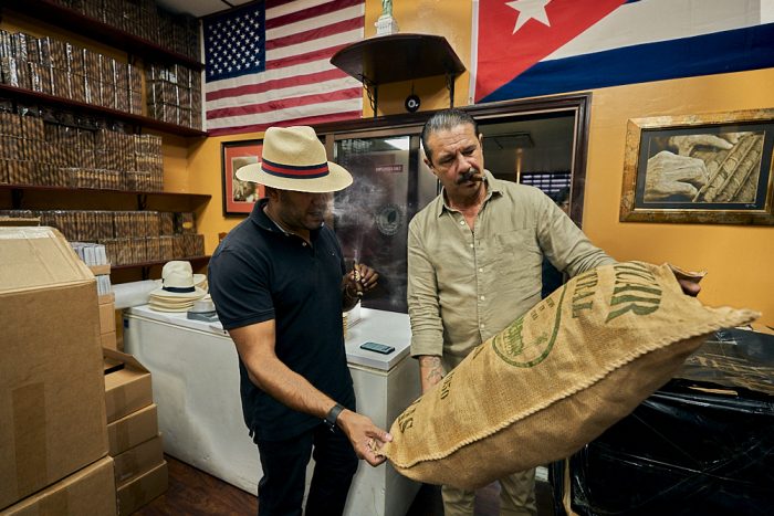 A horizontal photo of two men standing adjacent to each other while inspecting a large burlap sack of tobacco leaves. The man on the right holds the bag at a 45-degree angle, such that the man on his left can view the large printing on the surface. The man on the left reaches down with his right hand to hold the bottom of the bag to improve his angle of view. Both men smoke as they inspect the label, blowing puffs of dense white smoke. The man on the left wears a straw fedora had with a straight 3-inch brim and a red and black hat band. Above them on the back wall we see a United States flag, with the flag of Puerto Rico adjacent to the right. On another wall visible to the left, rows of cigars packaged in cellophane sit neatly stacked on dark wooden shelves.