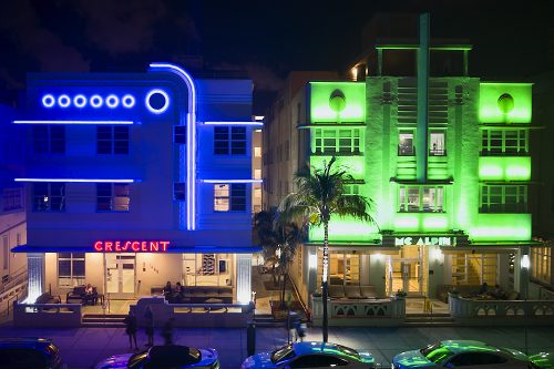 A horizontal drone photo on Ocean Drive in Miami’s South Beach at night, taken 30-feet in the air from the park across the street. A dark, deep purplish-blue light cloaks the three-story Crescent Hotel on the left—accentuated with blue neon art deco accent lights: Seven blue neon circles in a row hover just below the flat roofline. Horizontal blue neon lights line the tops of the windows on the second and third levels. On the right third of the façade, a pair of blue neon lights rise vertically from the portico to the roof, curving 90-degrees left over the blue circles. The Hotel's name, "Crescent," blazes in red neon art deco letters centered over the first-floor portico. Adjacent and to the right, Mc Alpin's three-story façade glows in a shimmering, amplified ultraviolet green. The illuminated 'Mc Alpin' sign, centered over the portico, blazes in an intense lime green over the entrance. Both façades look like art deco cubes: the dark blue Crescent on the left; bright green Mc Alpin on the right.