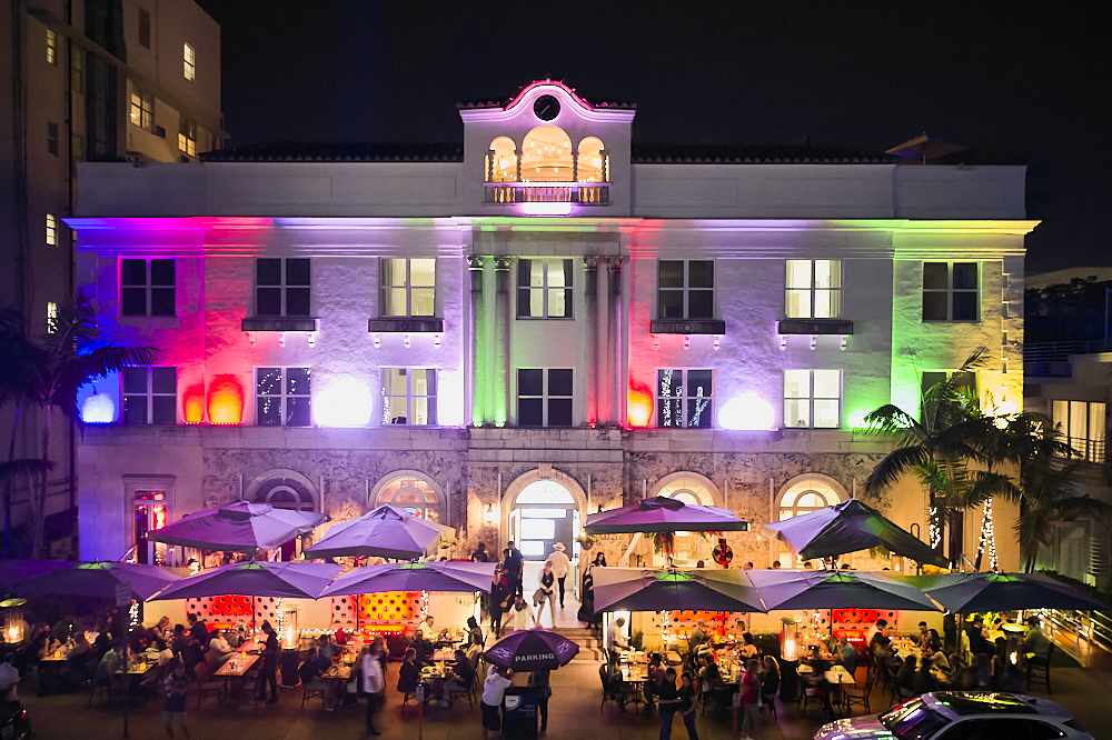 A horizontal drone photo on Ocean Drive in Miami’s South Beach at night. Taken 30-feet in the air from the park across the street, the composition looks directly at the center of Havana 1957, an authentic Cuban restaurant. Columns of multicolored lights between the windows on the second and third levels spotlight the building's long, white façade with pillars of intense blue, red, purple, green, and yellow. Ten square umbrellas line Ocean Drive, underneath which a hundred or more patrons dine alfresco. A man with a white shirt and white panama had stands at the entrance to the restaurant looking inside.