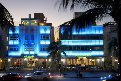 A horizontal drone photo on Ocean Drive in Miami’s South Beach at night. Taken from 30-feet in the air, the image looks directly at the Mc Alpin and Ocean Plaza Hotels from the park across the street. Bands of bright blue lights illuminate the facades of both buildings, in between layers of windows on each of the three levels. Palm frond silhouettes frame the left and right foreground of the image. A pale lavender-gray sky fades to twilight behind the buildings. Car headlights and taillights streak by horizontally on Ocean Drive, captured by the long shutter.