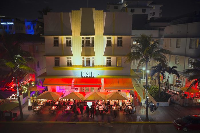 This night shot of the Leslie Hotel on Ocean Drive in Miami’s South Beach was taken from a drone hovering 40-feet across the street. Twin gold-lit vertical art deco fan facades reach from the second floor to the top of the roof. A red awning is brightly illuminated from the street level underneath, where tables are crowded with alfresco diners. A dark night sky and dimly lit street frame the building and outdoor dining scene top and bottom.