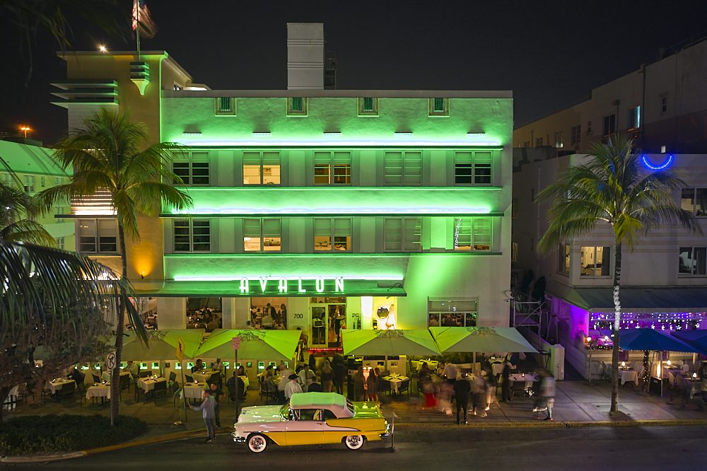 A horizontal drone photo on Ocean Drive in Miami’s South Beach at night. Taken from 30-feet in the air from the park across the street, the composition looks straight to the center of the Avalon Hotel. Bands of bright green neon lights stretch across each of the three-story balconies, in between a checkerboard of backlit and unlit windows. The hotel’s signature “Avalon” horizontal sign, in large art-deco font, illuminates the front of the building’s second story. Square yellow umbrellas line Ocean Drive where guests dine alfresco. A lone, white and yellow 57’ Chevy convertible, with 4-inch whitewall tires and lots of ‘attitude,’ sits proudly, parked on the street. The polished hood and trunk of the Chevy glistens underneath the green lights of the hotel’s façade.