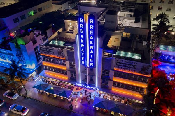 A horizontal drone photo on Ocean Drive in Miami’s South Beach at night. Taken from 150-feet in the air, the image looks down to the Breakwater Hotel from the park across the street. The Breakwater’s long vertical blue neon sign rises from the second story, reaching 50-feet or more over the top of building’s 4-story frame. Two mint green colored swimming pools hug the front edge of the roof on either side of the large vertical sign. Orange neon horizontal lights outline the second, third, and fourth floors. On the street level far below, square awnings line Ocean Drive, where guests dine alfresco.