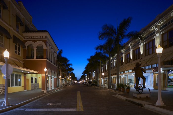 Looking northwest from the center of brick-paved First Street in downtown Fort Myers, the faint light of an early dawn glows blue at the vanishing point on the horizon, brightening the dark sky overhead. Streetlamps illuminate the 2-story building fronts while 30-foot palms lining the street are silhouetted by dawn on the horizon. The street is empty and quiet except for metal sidewalk street art of a man balancing on a unicycle.