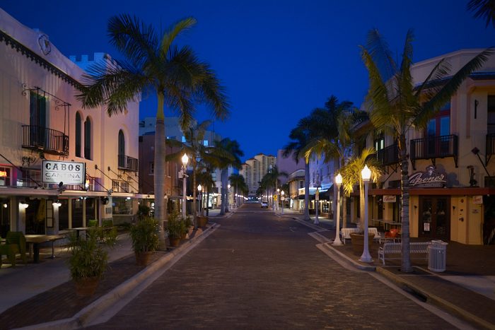 Looking southwest and down the street from the center of brick-paved First Street in downtown Fort Myers, both curbs diminish toward the vanishing point almost 1-mile distant. Streetlamps in front of 30-foot palm trees line the avenue and illuminate Capone’s Coal Fired Pizza and Cabos Cantina in the foreground. The street is completely empty as the black sky overhead slowly fades to dawn. At the end of the street, and in the center of the photograph, High Point Place, a 33-story high-rise condominium, faintly catches early rays of the rising sun.