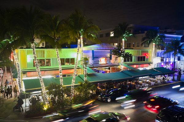 In a night drone shot, the drone hovers 30-feet on the corner of Ocean Drive and Ninth Street overlooking Mango’s Tropical Café in South Beach, Miami. White Italian miniature lights decorate the trunks of 40-foot palm trees for Christmas in front of the restaurant. The palm treetops frame the top of the image. The second story façade is brightly lit in a bright greens and blues. Patrons sit outside underneath a dark green awning that runs the length of the restaurant. Headlights and taillights of passing cars blur in motion as they pass in front.