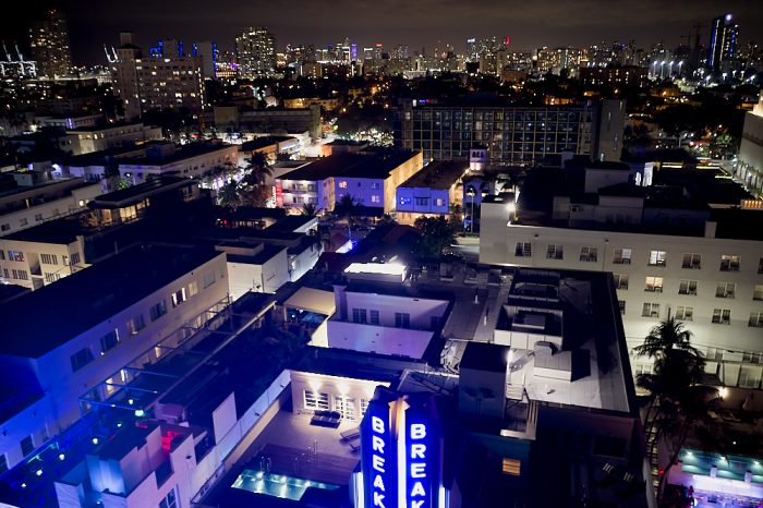 A drone, hovering 100-feet or more at night, looks west over the Breakwater Hotel’s blue vertical backlit sign and roof, at the bottom of the image, to Miami’s skyline over 3-miles away. Buildings and rooftops lit from the outside—many with illuminated windows—compose the lower 2/3s of this landscape image. Just beneath the black sky at the top of the image, Miami’s high-rise architecture fills the horizon with hundreds of tiny neon and window lights from the buildings on the skyline.