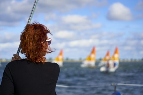 Sitting on the observation catamaran I look abeam over the shoulder of a woman with deep red hair and a dark black sweater who is on the left one-third of the image. The right side of her face turns toward the center of the image as she watches a sailboat race. We see the edge of her red framed sunglasses and white temple arm extending back and wrapping over her right ear. Her red hair blows away from me, onto her face. The center and right one-third of the image includes four Hobie 16 sailboats with white and orange sails, racing in the Hobie 16 World race off of Captiva Island, Florida, under a white cloud and bright blue sky. The sailboats and sky are heavily blurred because the image focuses on the woman.