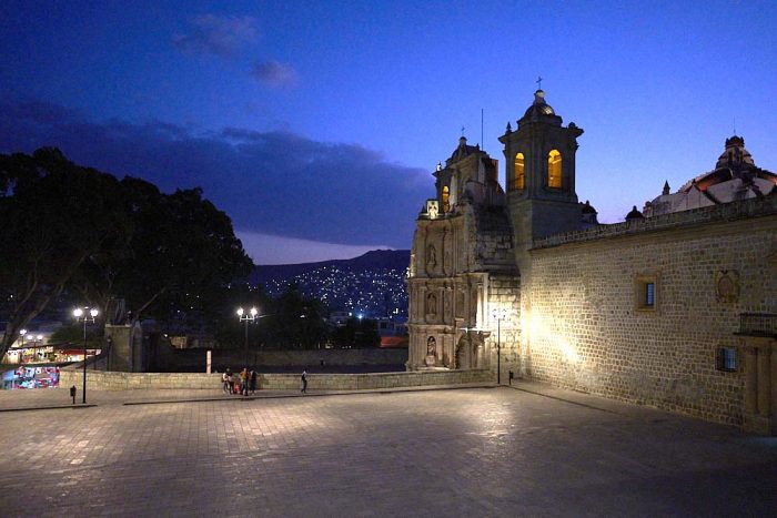 Overlooking the Plaza de la Danza with the Basilica of Our Lady of Solitude on the right side of the image. A dark blue twilight sky frames the silhouetted trees in the back of the plaza and twinkling lights on the nearby mountain in the background. Young friends gather to talk on the far side of the plaza underneath the lamp light. Photo is taken from Avenue Jose Maria Morelos.