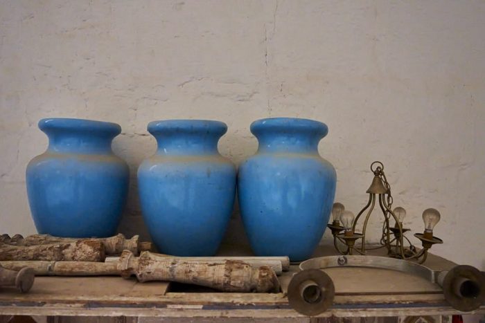 Horizontal photo of four large, light blue ceramic urns, 18-inches at the center diameter, that appear to have been abandoned in the church of Saint John the Baptist, located in San Juan Bautista Coixtlahuaca, Mexico. The dusty urns lean against a stucco wall, askew, on a table along with several discarded candleholders and an old hanging chain light fixture with four dirty bare lightbulbs.